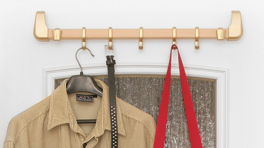 Obešalnik_nature_ZL_na vratih Zlati obešalnik nature line na belih vratih, na katerega je obešena torbica, pas in srajca.