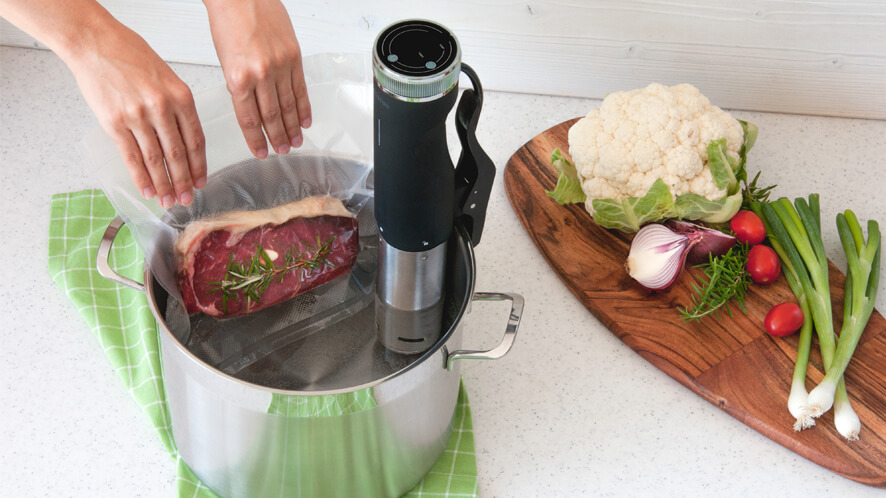 Sous Vide SVC100_polaganje vrečke v lonec Polaganje zavakuumiranega steaka v lonec z vodo na katerem je pripet počasni kuhalnik Status. Zraven še cvetača in čebula na leseni deski za rezanje.