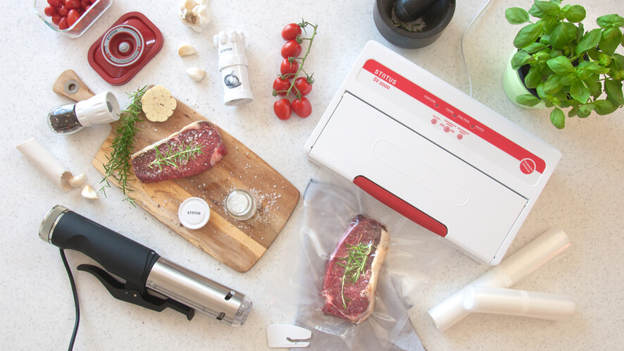 Sous Vide SVC100_SV2000 Počasni kuhalnik, vakuumski aparat z zavakuumiranim mesom, deska za rezanje in meso, paradižnik na belem pultu.